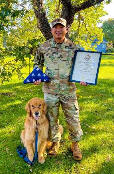 A veteran dressed in U.S. Army fatigues stands with a golden retriever by his side. The man holds his retirement certificate in one hand and, in the other, an American flag folded into a triangle.
