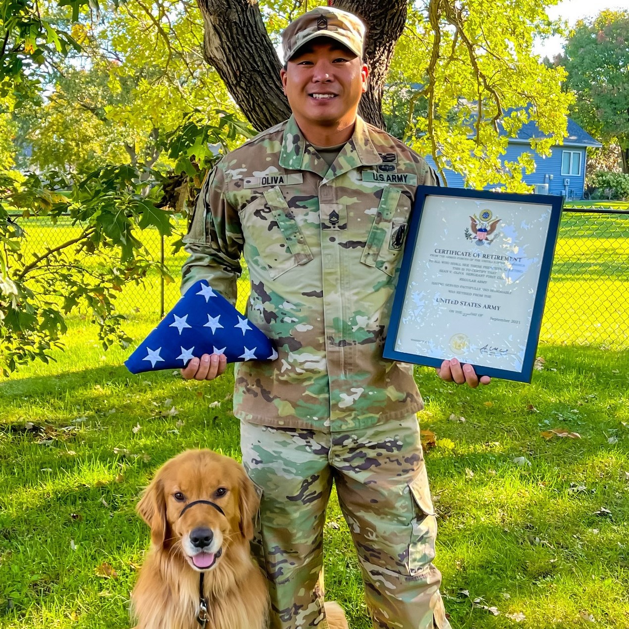 A veteran dressed in U.S. Army fatigues stands with a golden retriever by his side. The man holds his retirement certificate in one hand and, in the other, an American flag folded into a triangle.