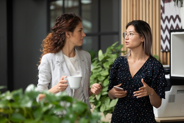 Two women, one white and one Asian, chat while on a coffee break from work, where they help to build a culture of equity.