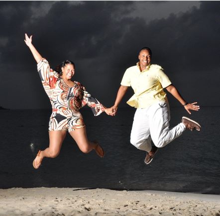 Two smiling Black women on a beach at nighttime, holding hands and leaping up in the air with happiness.